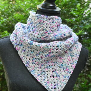 A textured, triangular crocheted cowl or small shawl is displayed outdoors on a black mannequin torso against a soft-focus background of green foliage. The fabric features a repetitive openwork granny-style stitch pattern made from a variegated yarn blending light grey, blue, pink, and purple hues. The accessory wraps snugly around the neck and drapes down into a central V-shape point in the front.