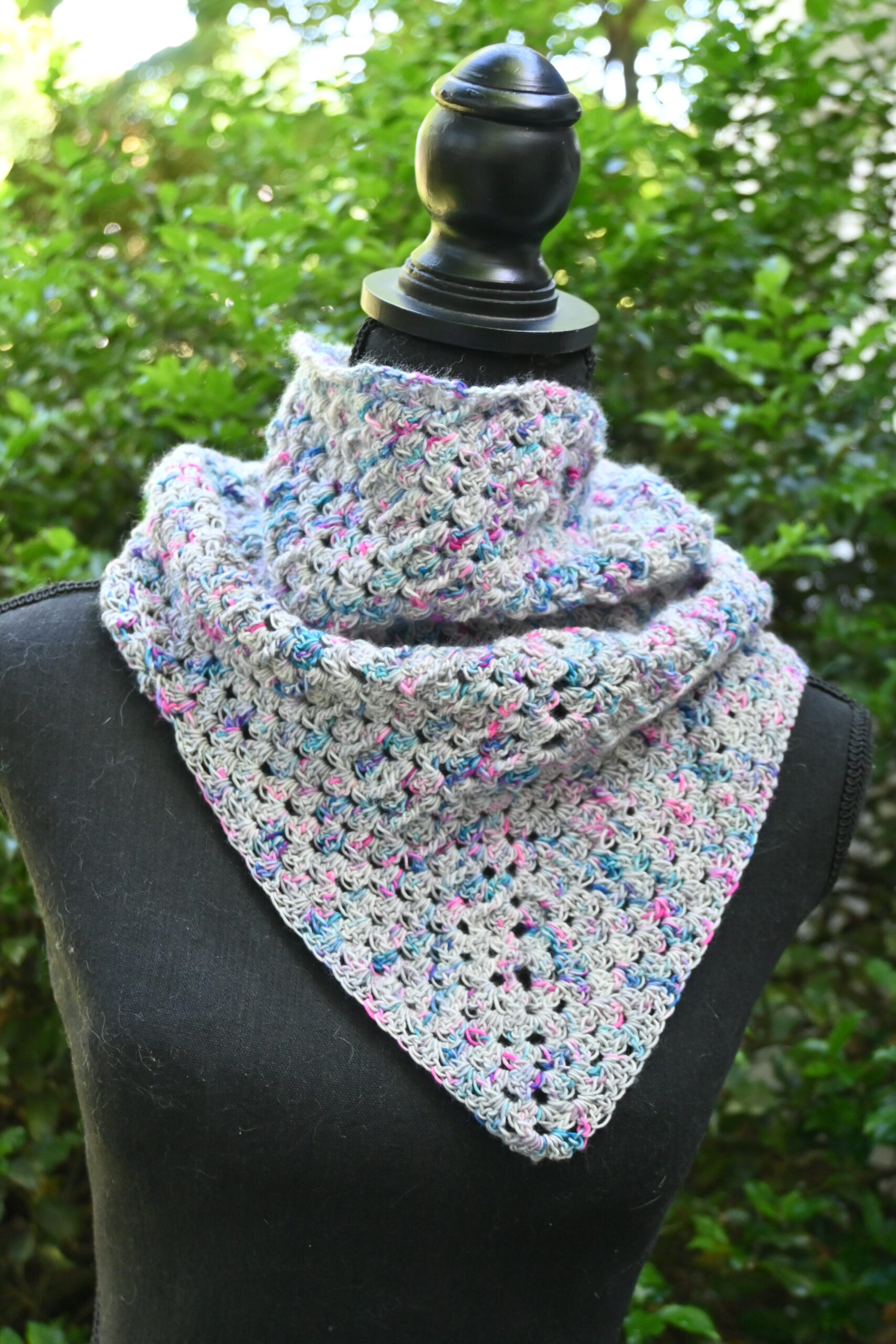 A textured, triangular crocheted cowl or small shawl is displayed outdoors on a black mannequin torso against a soft-focus background of green foliage. The fabric features a repetitive openwork granny-style stitch pattern made from a variegated yarn blending light grey, blue, pink, and purple hues. The accessory wraps snugly around the neck and drapes down into a central V-shape point in the front.
