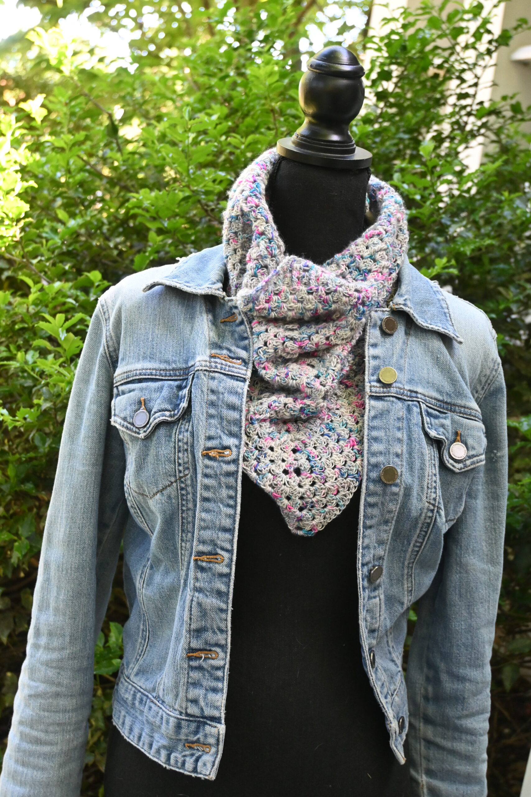 A close-up shot shows a textured, multicolored crocheted cowl or keyhole scarf styled around the neck of a black mannequin torso. The mannequin wears a classic light-wash denim jacket, which is left unbuttoned to showcase the drape of the scarf. The crocheted fabric features a speckled blend of light blue, pink, cream, and grey yarn. In the top-right corner, a small white square logo reads "First Byte Designs."