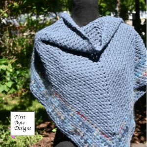 A triangular, textured blue crocheted shawl is displayed outdoors on a black mannequin torso against a backdrop of green foliage. The main body of the shawl is a solid dusty blue color with an openwork, patterned stitch, while the bottom border features a multicolored flecked pattern with subtle hints of red and cream. A white square logo reading "First Byte Designs" is visible in the bottom left corner.