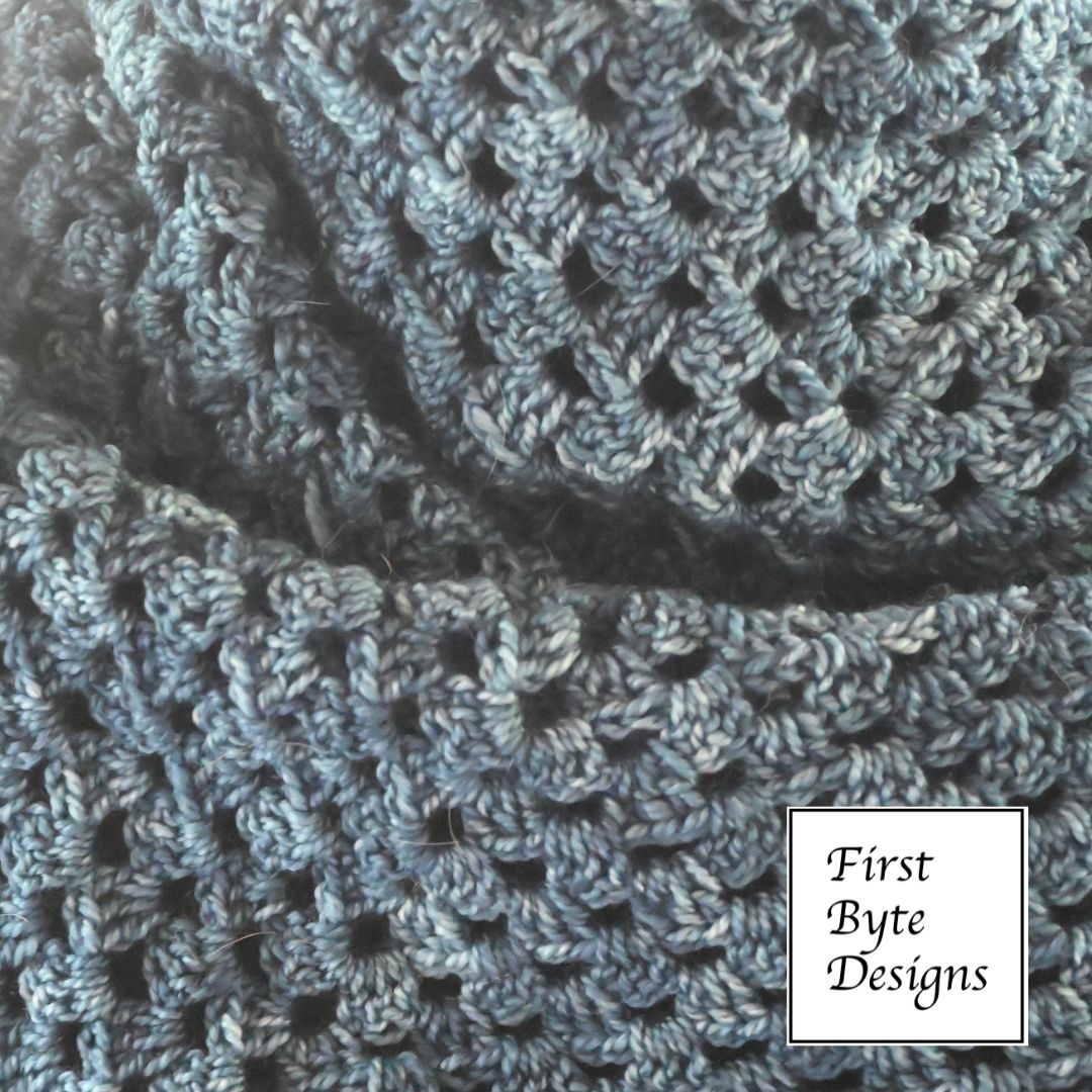 A close-up photograph displays a textured, blue-grey crocheted fabric with an openwork granny square pattern. The fabric is softly folded, creating shadows that emphasize the intricate yarn loops and repeating mesh-like spaces. In the bottom right corner, a white square logo with a thin black border features the text "First Byte Designs" in a classic serif font.