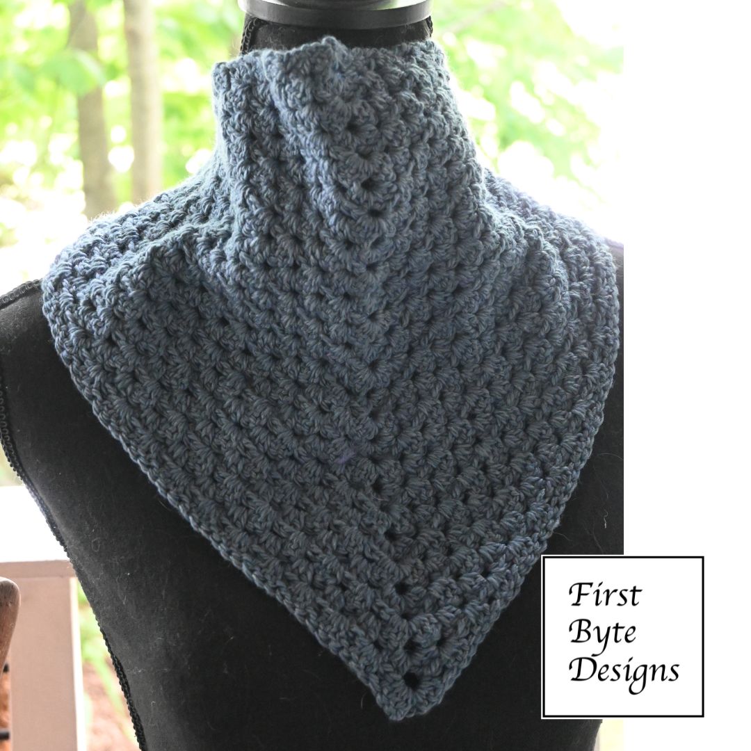 A steel-blue crocheted cowl or bandanna-style scarf is modeled on a black mannequin torso. The garment fits closely around the neck like a high collar and points downward in a sharp triangle over the chest, showcasing a dense, repeating granny stitch texture. In the lower right corner, a white square logo reads "First Byte Designs"