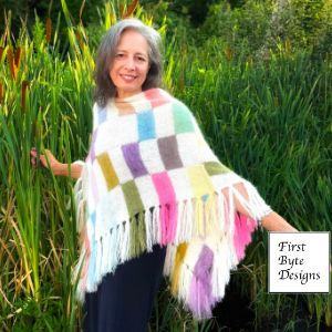 A woman smiling outdoors wearing a handmade, multi-colored patchwork knit poncho with white borders and long fringe detailing at the hem